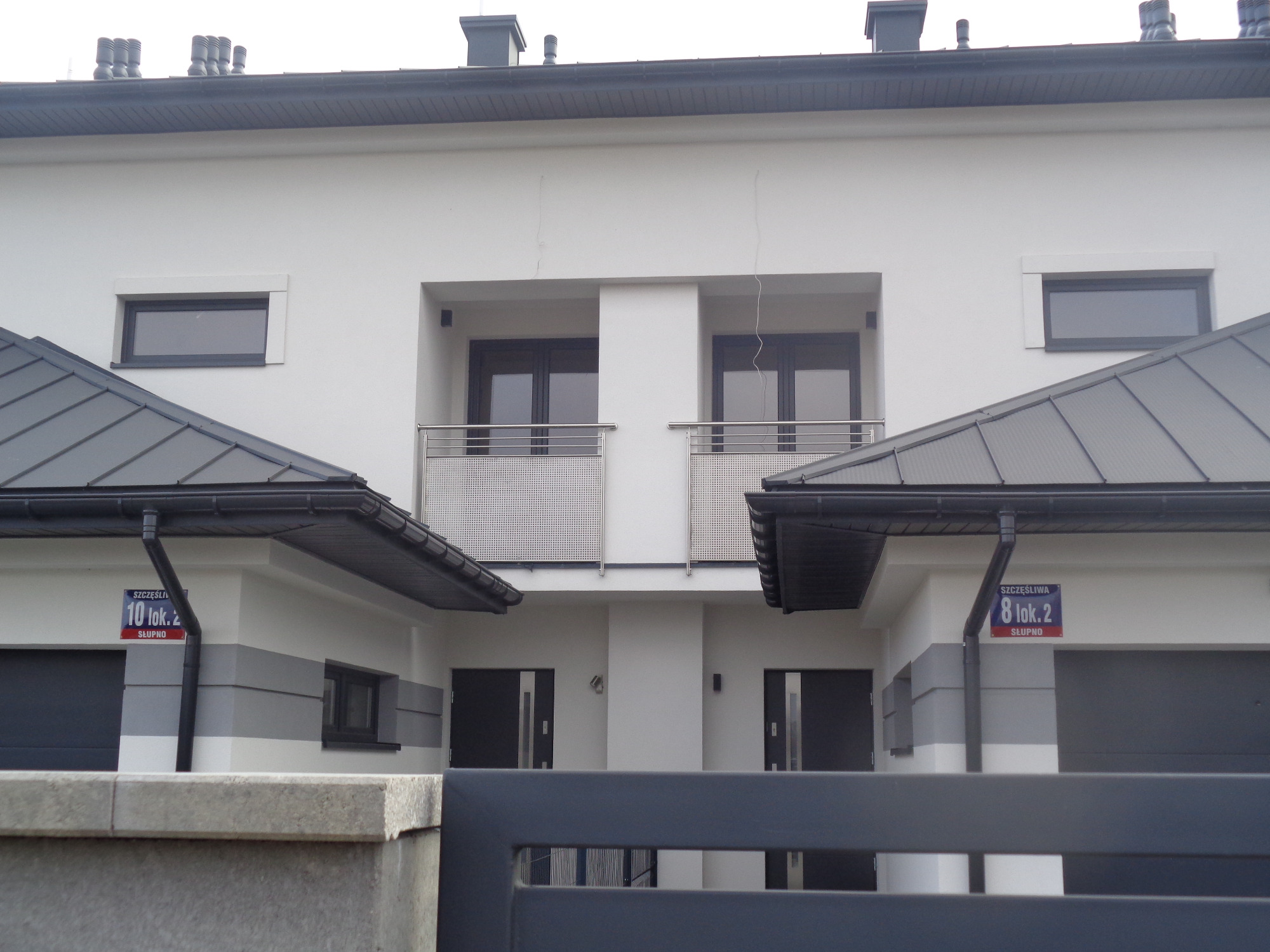 Bliźniak z balkonami zabezpieczonymi metalowymi balustradami, widok zza ogrodzenia.