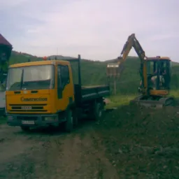 Żółty samochód ciężarowy marki Iveco z napisem 'Construction' zaparkowany obok małej koparki podczas prac ziemnych na nierównym terenie.