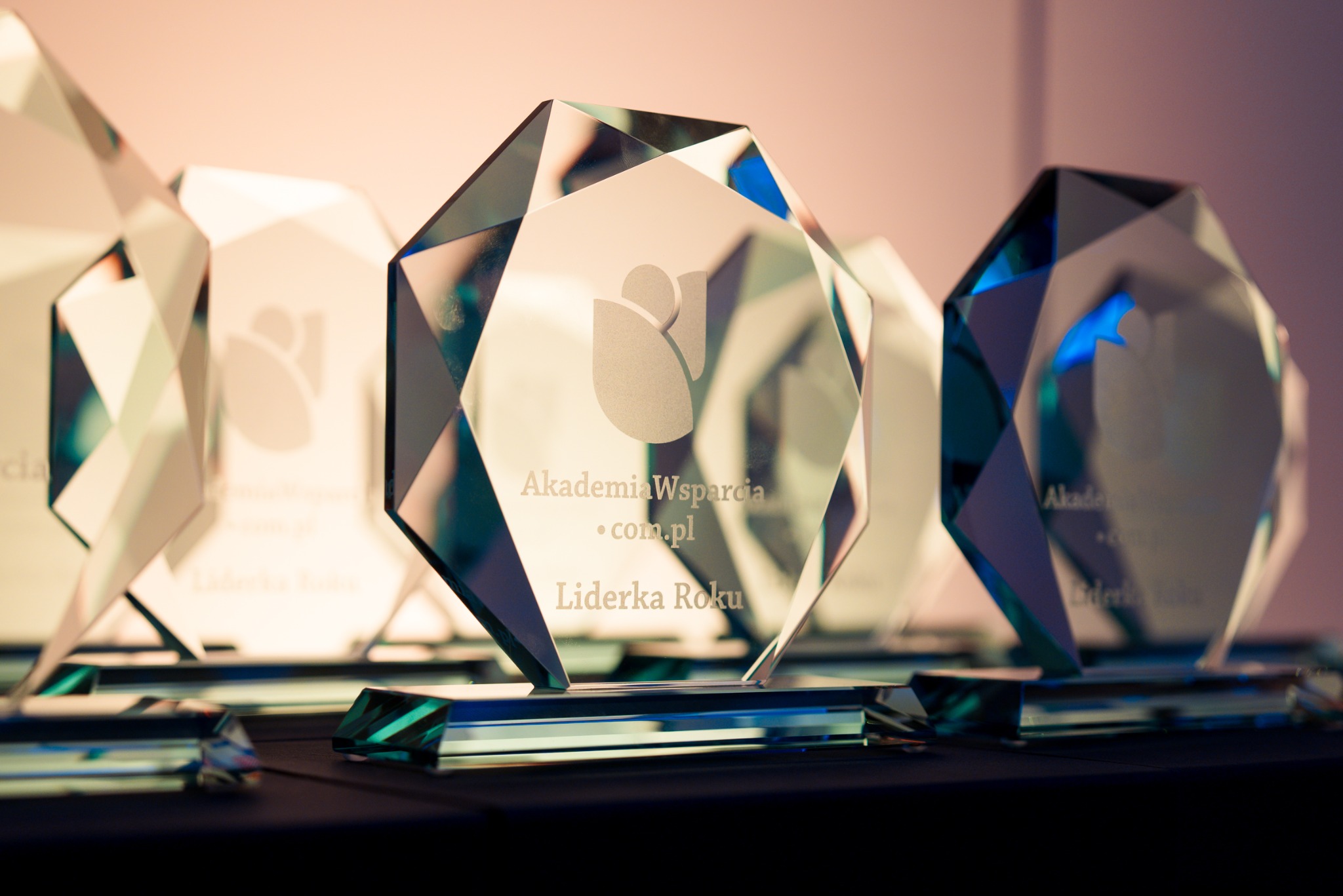 Szklane statuetki nagrody 'Liderka Roku' dla Akademii Wsparcia, ustawione rzędem na ciemnym blacie, z logo i nazwą firmy wygrawerowaną na powierzchni.