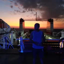 Mężczyzna na pokładzie promu na tle zachodzącego słońca. Widoczne elementy konstrukcji promu i fragment morza. Scena oświetlona sztucznym, niebieskim światłem.