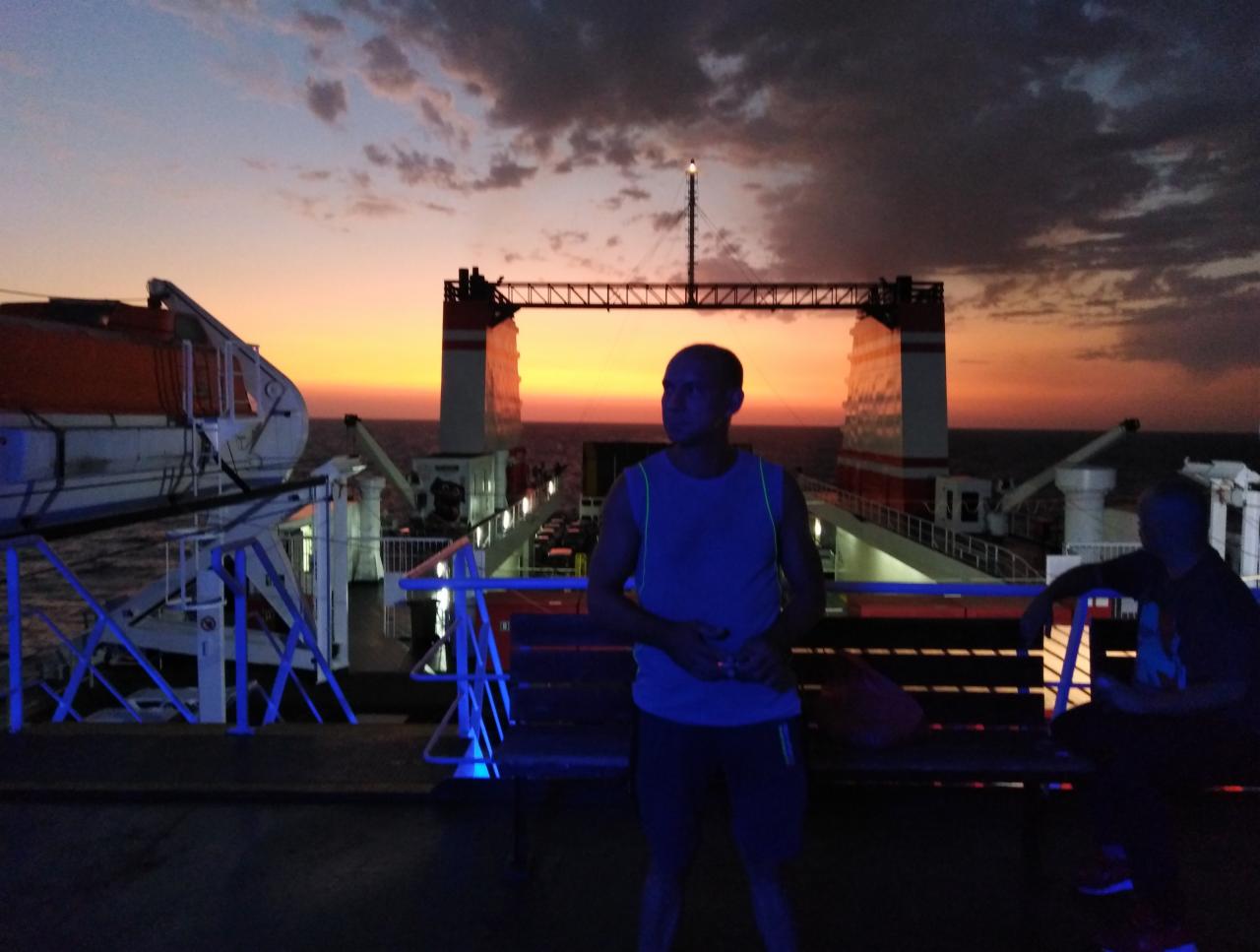 Mężczyzna na pokładzie promu na tle zachodzącego słońca. Widoczne elementy konstrukcji promu i fragment morza. Scena oświetlona sztucznym, niebieskim światłem.