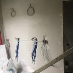 Ściana w trakcie prac instalacyjnych: widoczne wystające rury wodne, kable elektryczne w puszkach, surowy tynk i worki z materiałami budowlanymi na podłodze.