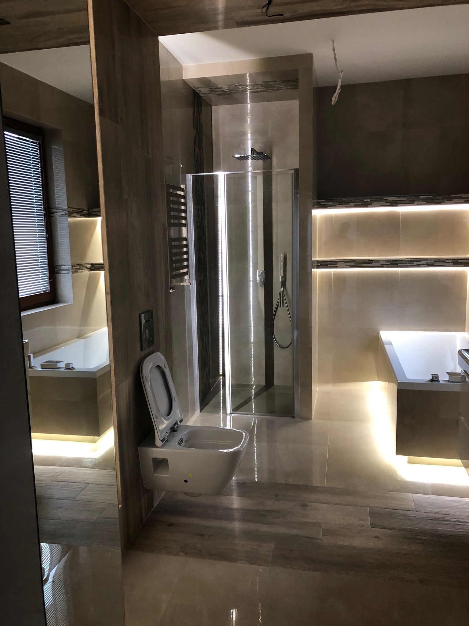 Nowoczesna łazienka z kabiną prysznicową, podwieszaną toaletą i wanną z podświetleniem LED, wykończona w beżowych płytkach imitujących drewno, z widocznymi elementami instalacji elektrycznej...