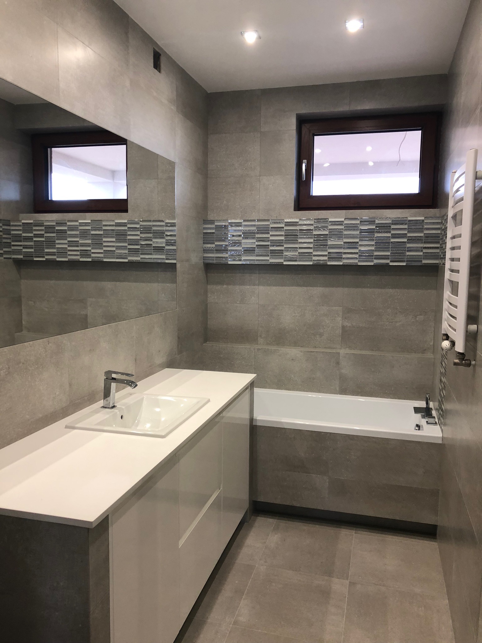 Nowoczesna łazienka z szarymi płytkami, białymi meblami łazienkowymi, wanną i oknem z brązową ramą. Nad wanną i umywalką dekoracyjny pas mozaiki. Na ścianie zamontowany grzejnik drabinkowy.