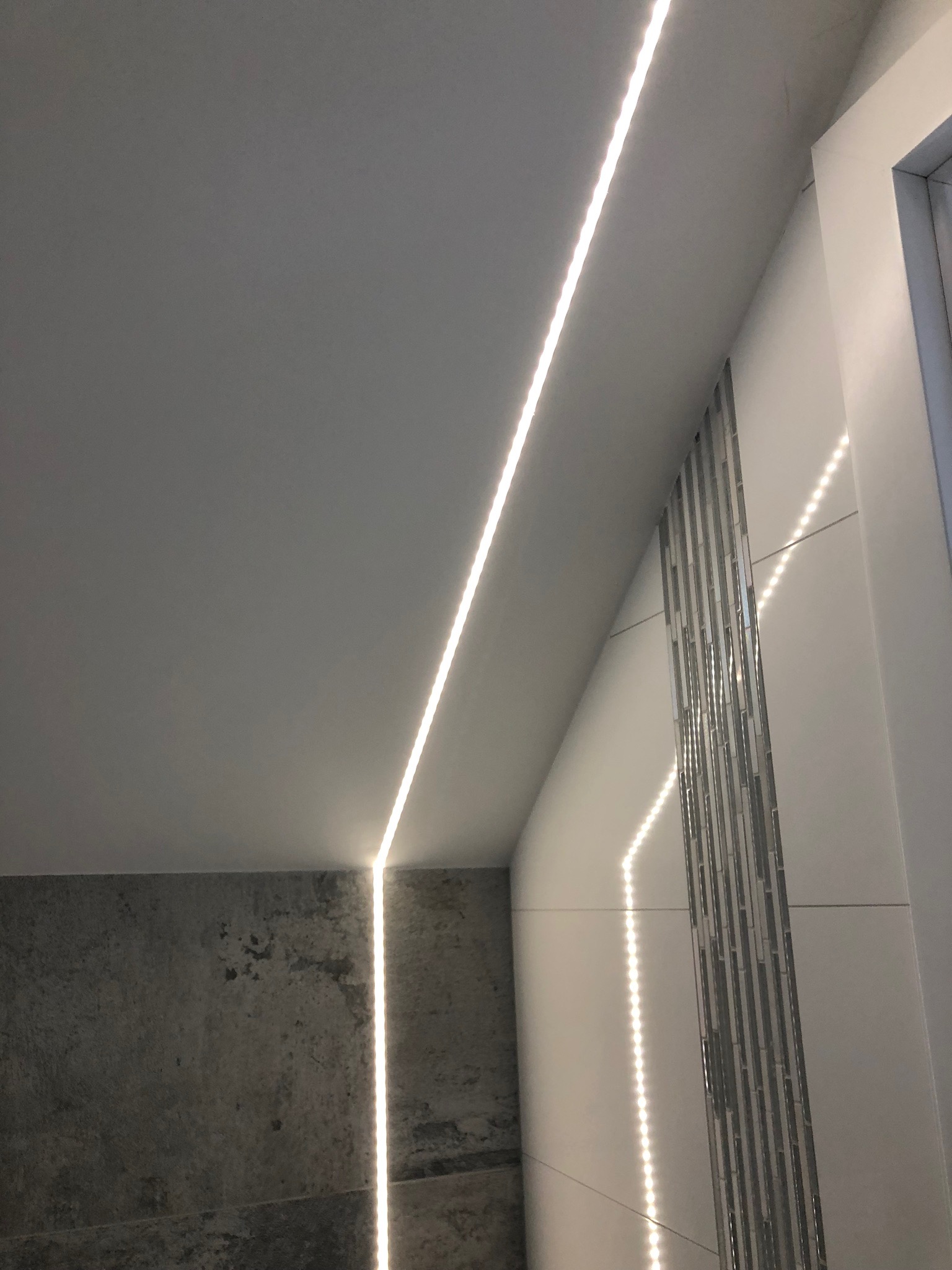 Nowoczesna łazienka z podświetleniem LED wzdłuż sufitu i ściany, akcentująca szarą teksturę płytek i dekoracyjne panele ścienne.