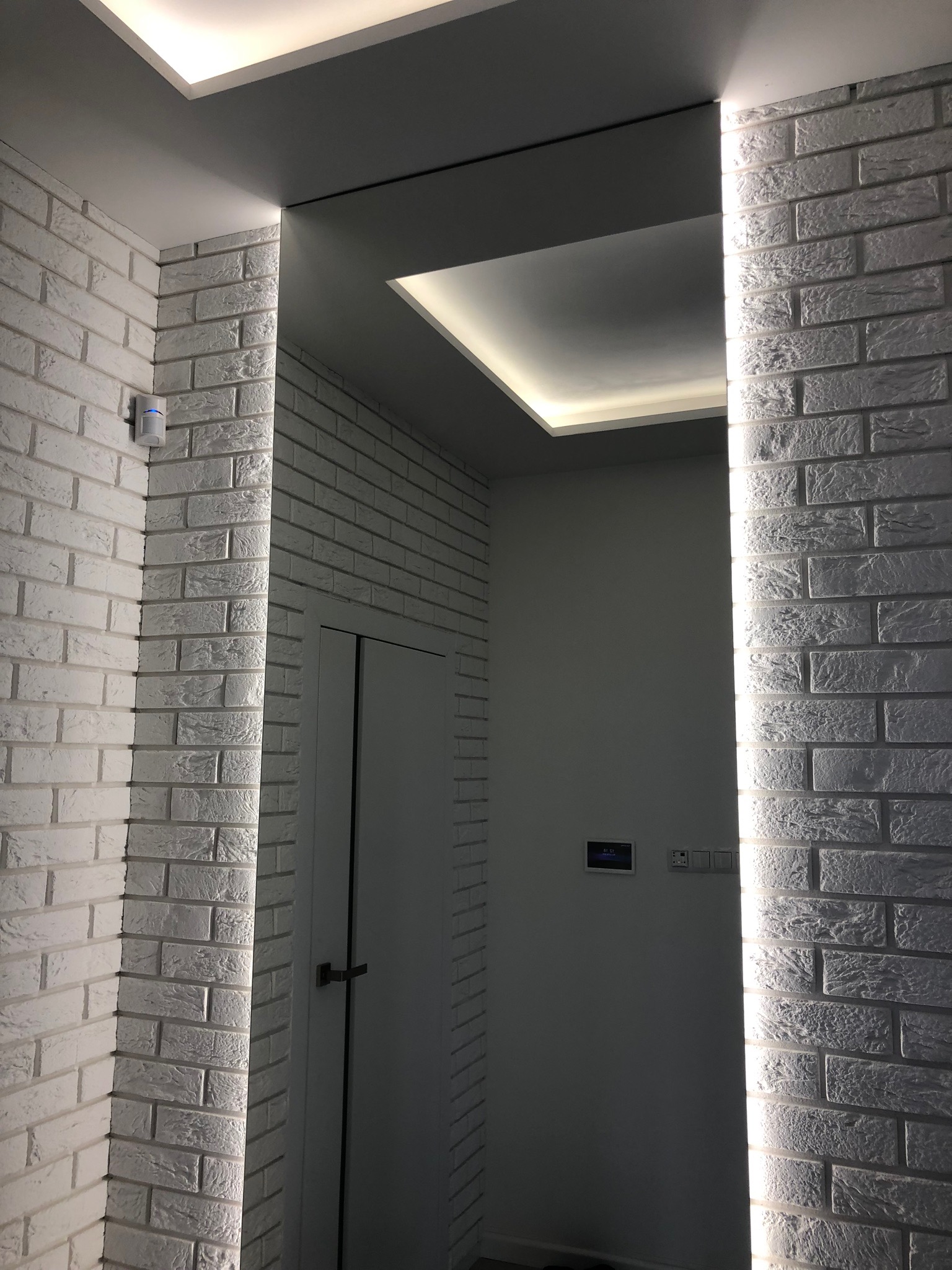 Korytarz wykończony białą cegłą z podświetleniem LED, widoczny czujnik ruchu i panel sterowania na ścianie, drzwi w tle.