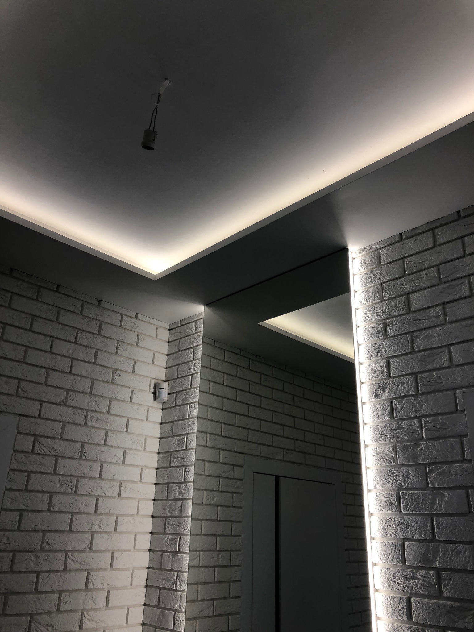 Nowoczesne oświetlenie LED w suficie podwieszanym z szarą wnęką, biała cegła na ścianach, widoczna oprawka żarówki na kablu.