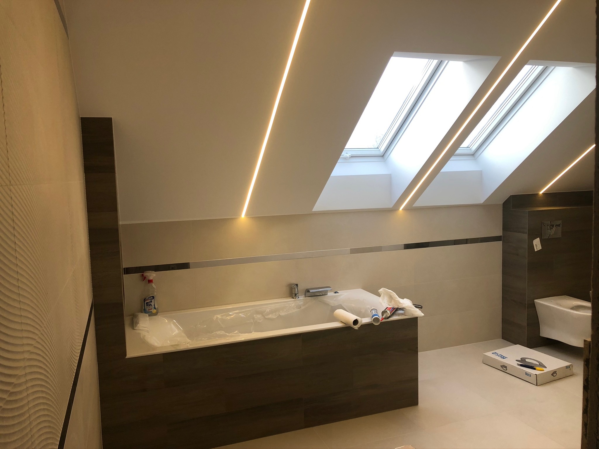 Łazienka w trakcie remontu: zabezpieczona folią wanna, widoczne narzędzia i materiały budowlane, oświetlenie LED w suficie skośnym z oknami dachowymi.
