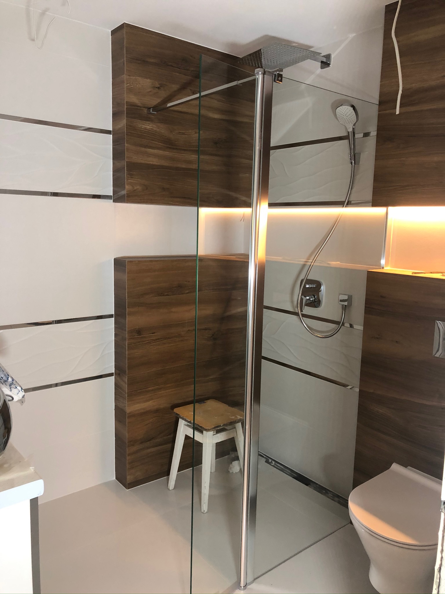 Nowoczesna kabina prysznicowa walk-in z drewnopodobnymi elementami wykończeniowymi, deszczownicą i oświetleniem LED, widoczny fragment toalety i roboczy stołek.