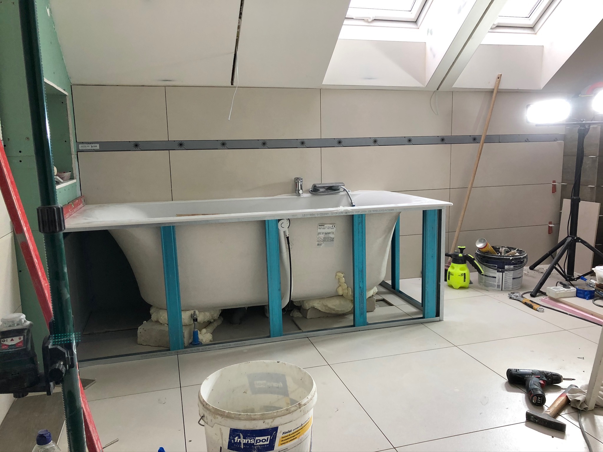 Montaż wanny w obudowie z metalowych profili, z widocznym ociepleniem pianką montażową, na tle wykończonych płytek ściennych z dekoracyjnym pasem, narzędzia i materiały budowlane na podłodze.