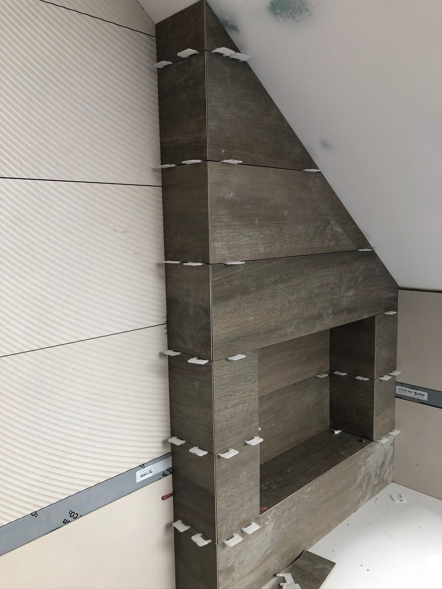 Montaż płytek imitujących drewno wokół kominka w trakcie remontu, z użyciem poziomicy i krzyżyków dystansowych, widoczne białe płytki z falistym wzorem na ścianie obok.