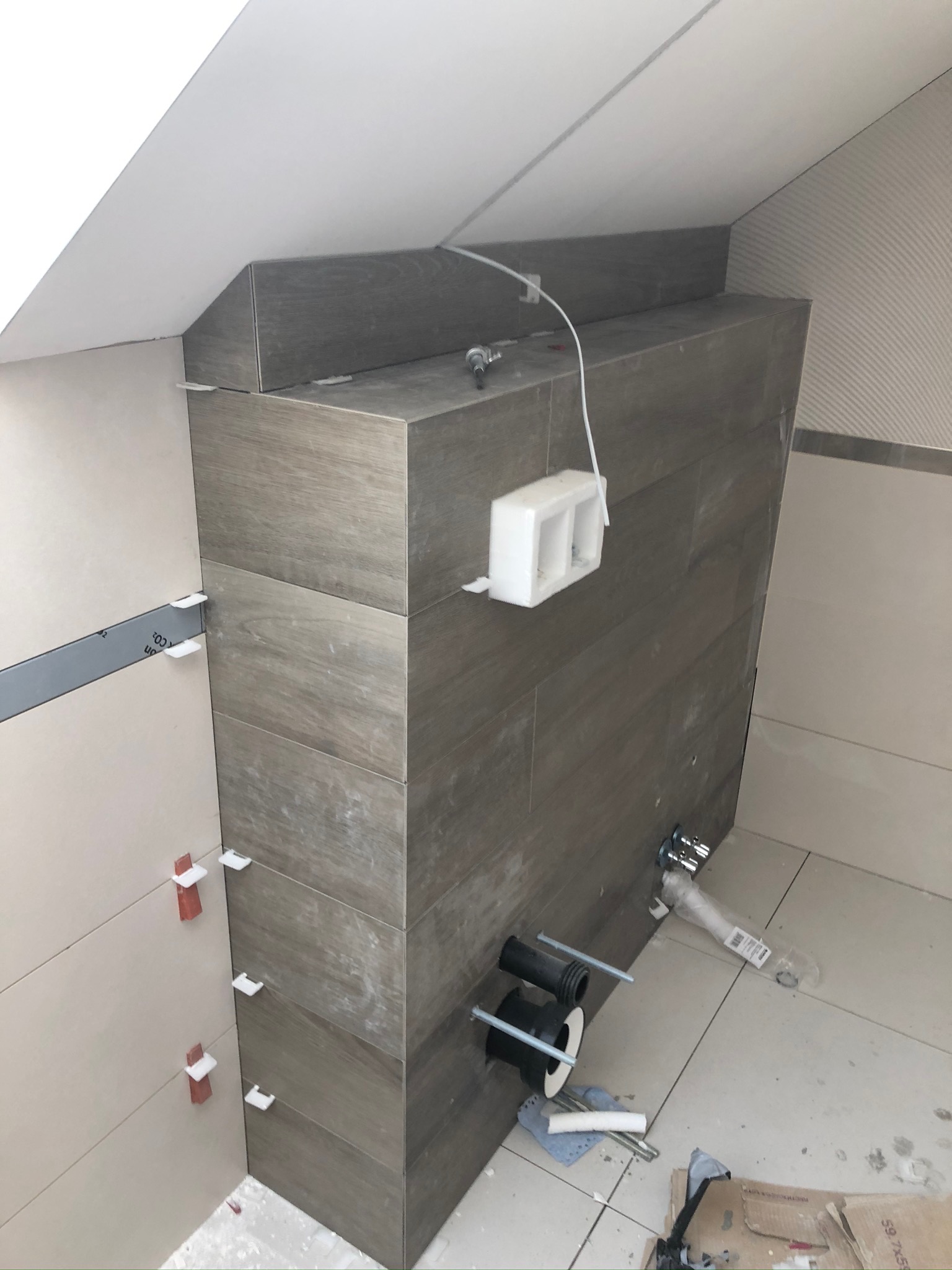 Montaż podtynkowej instalacji sanitarnej w łazience na poddaszu, widoczne płytki imitujące drewno, stelaż podtynkowy, rury kanalizacyjne, oraz uchwyty dystansowe do płytek.