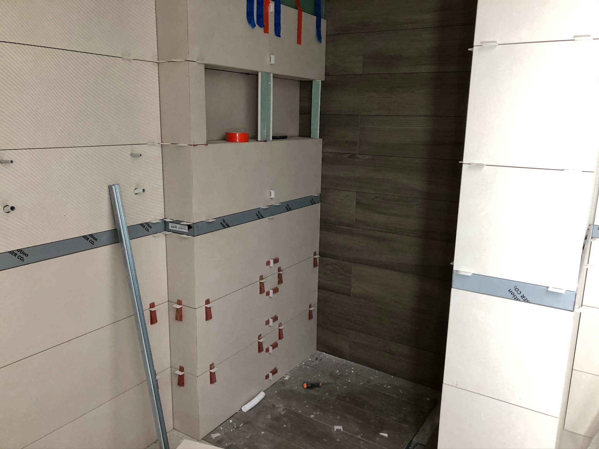 Montaż jasnych, podłużnych płytek w łazience z widocznymi krzyżykami dystansowymi, aluminiową listwą i poziomnicą, wnęka z elementami montażowymi, obok ściana z ciemnymi płytkami imitującymi drewno.