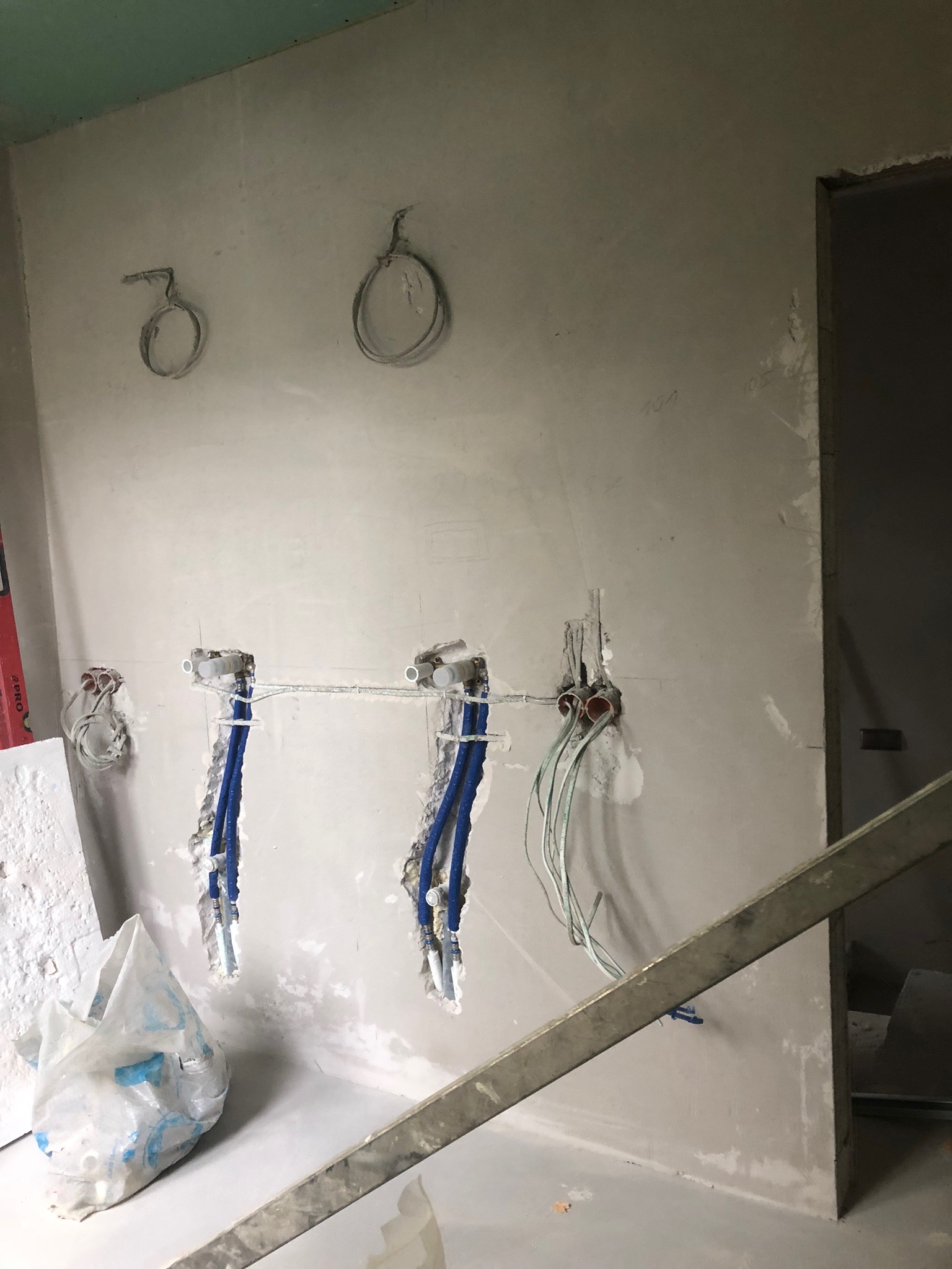 Ściana w trakcie prac instalacyjnych: widoczne wystające rury wodne, kable elektryczne w puszkach, surowy tynk i worki z materiałami budowlanymi na podłodze.