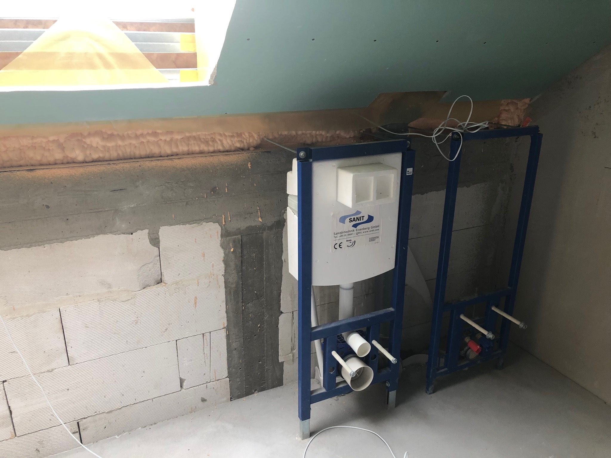 Stelaż podtynkowy WC marki Sanit zamontowany w surowym wnętrzu łazienki na poddaszu, widoczne białe bloczki betonowe, ocieplenie pianką montażową przy oknie dachowym oraz instalacje hydrauliczne.