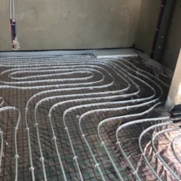 Białe rury układane w pętlach na folii z czerwoną siatką, mocowane plastikowymi klipsami, przygotowane do zalania wylewką, widoczne fragmenty ścian w stanie surowym.