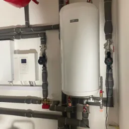 Biały zasobnik ciepłej wody użytkowej Nibe UKV z szarą izolacją rur, zaworami i sterownikiem Nibe SMO 20 na białej ścianie w kotłowni.