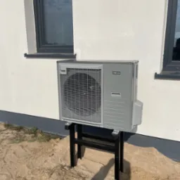 Szara pompa ciepła NIBE AMS20 zamontowana na czarnym stojaku przy białej ścianie budynku z dwoma oknami. Ujęcie z dołu, widoczne podłoże z piasku.