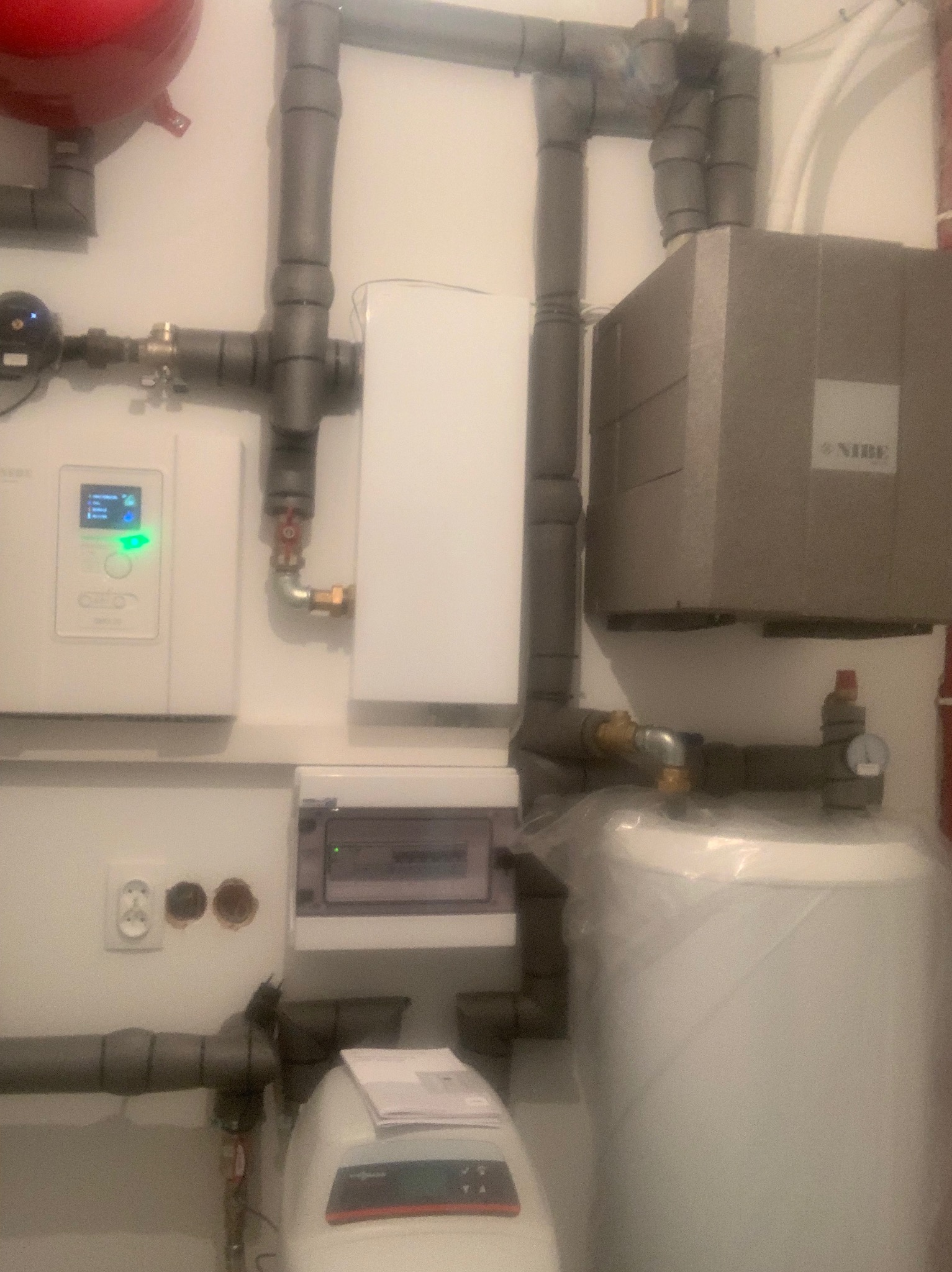 Instalacja grzewcza z kotłem, zbiornikiem i rurami w izolacji, widoczny panel sterowania i rozdzielnica elektryczna, częściowo zakryty zbiornik owinięty folią ochronną.