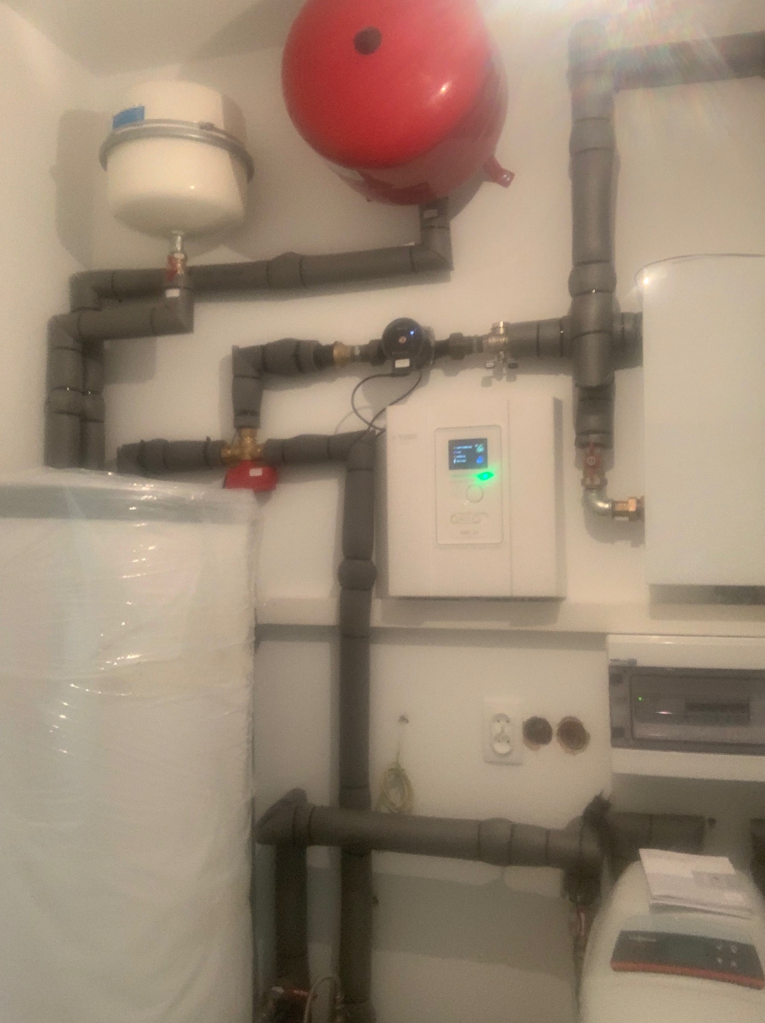 Złożona instalacja grzewcza z widocznym zbiornikiem wyrównawczym, naczyniem przeponowym, rurami izolowanymi termicznie oraz panelem sterowania z zielonym wyświetlaczem, zamontowana na białej ścianie.