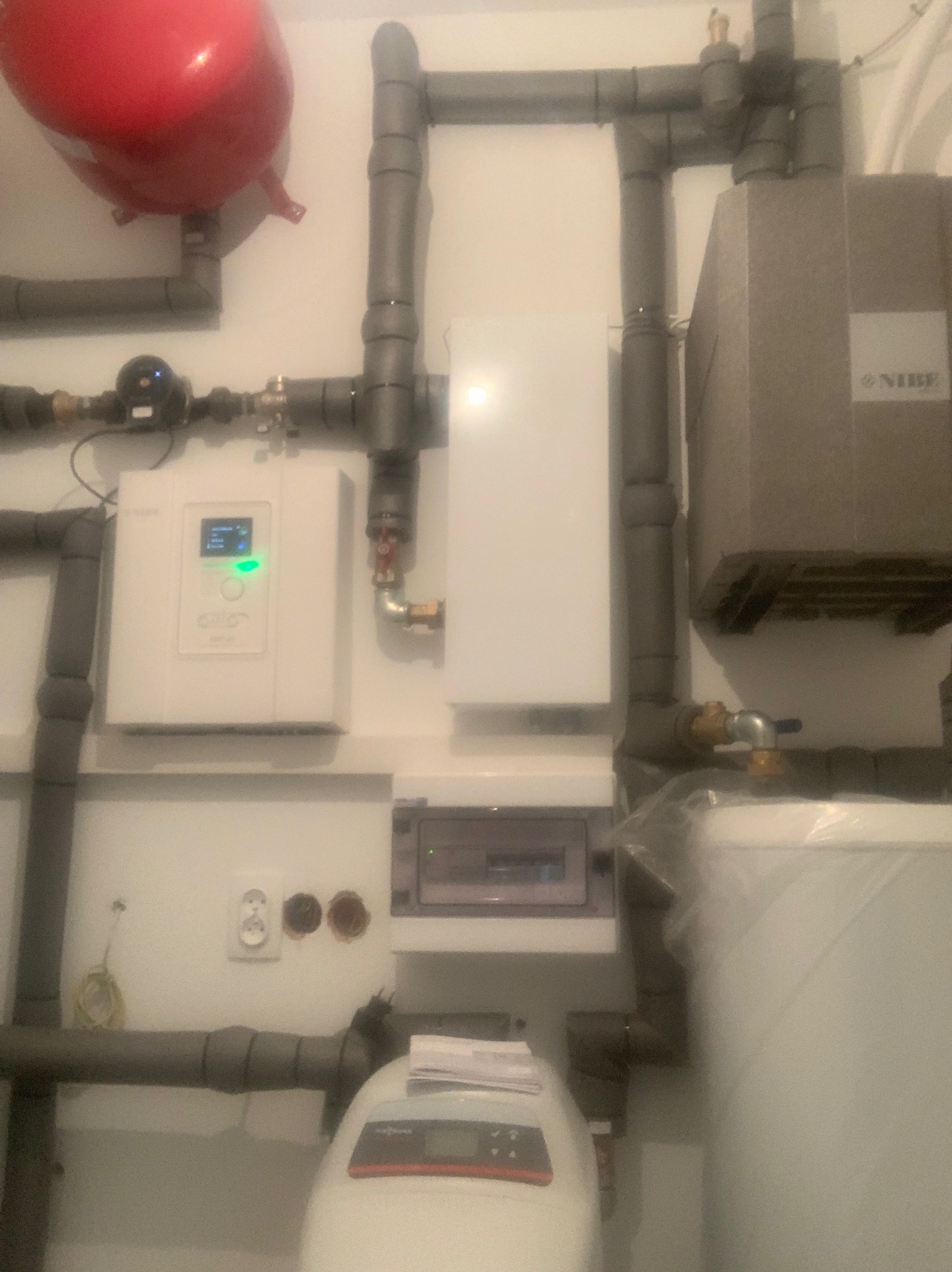 Instalacja grzewcza z czerwonym zbiornikiem ciśnieniowym, białym panelem sterowania z zieloną diodą, szarą izolacją na rurach i skrzynką elektryczną na białej ścianie.