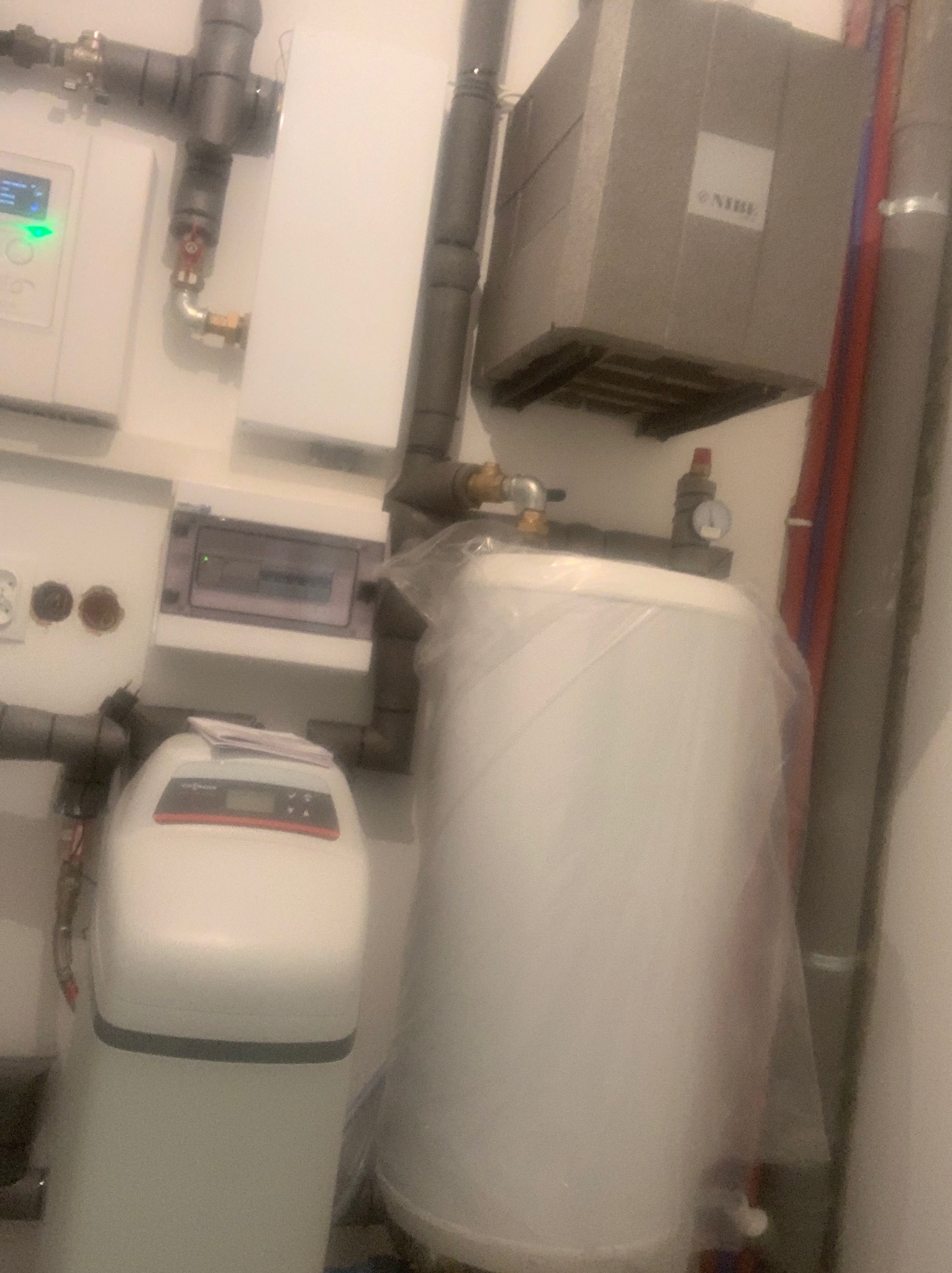 Instalacja kotła grzewczego i zmiękczacza wody w kotłowni, widoczne rury, zawory, manometr i elektronika sterująca, kocioł jeszcze w folii ochronnej.