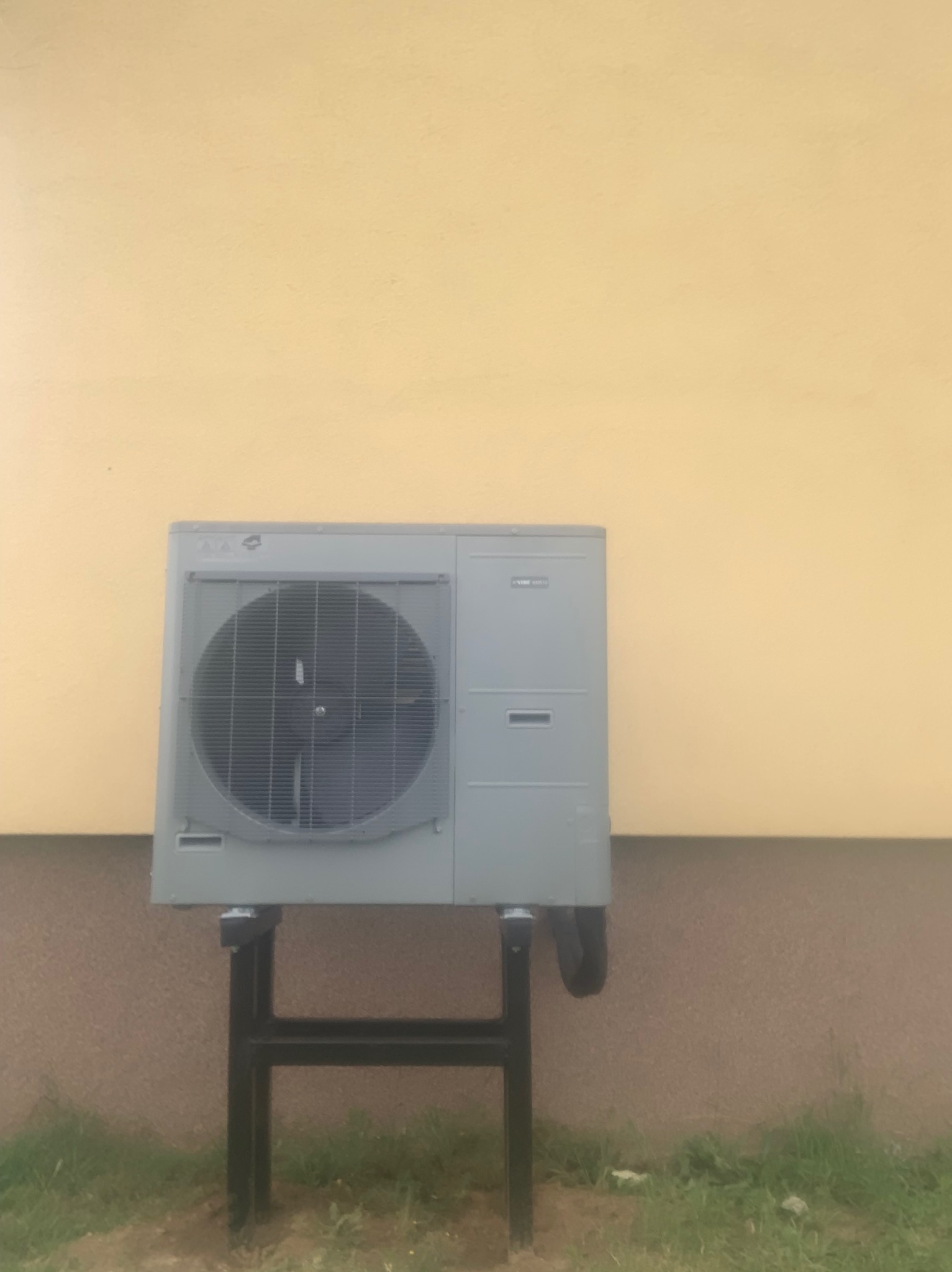 Zewnętrzna jednostka klimatyzacji zamontowana na czarnej, metalowej konstrukcji wsporczej przy ścianie budynku o żółtej górnej i brązowej dolnej części.