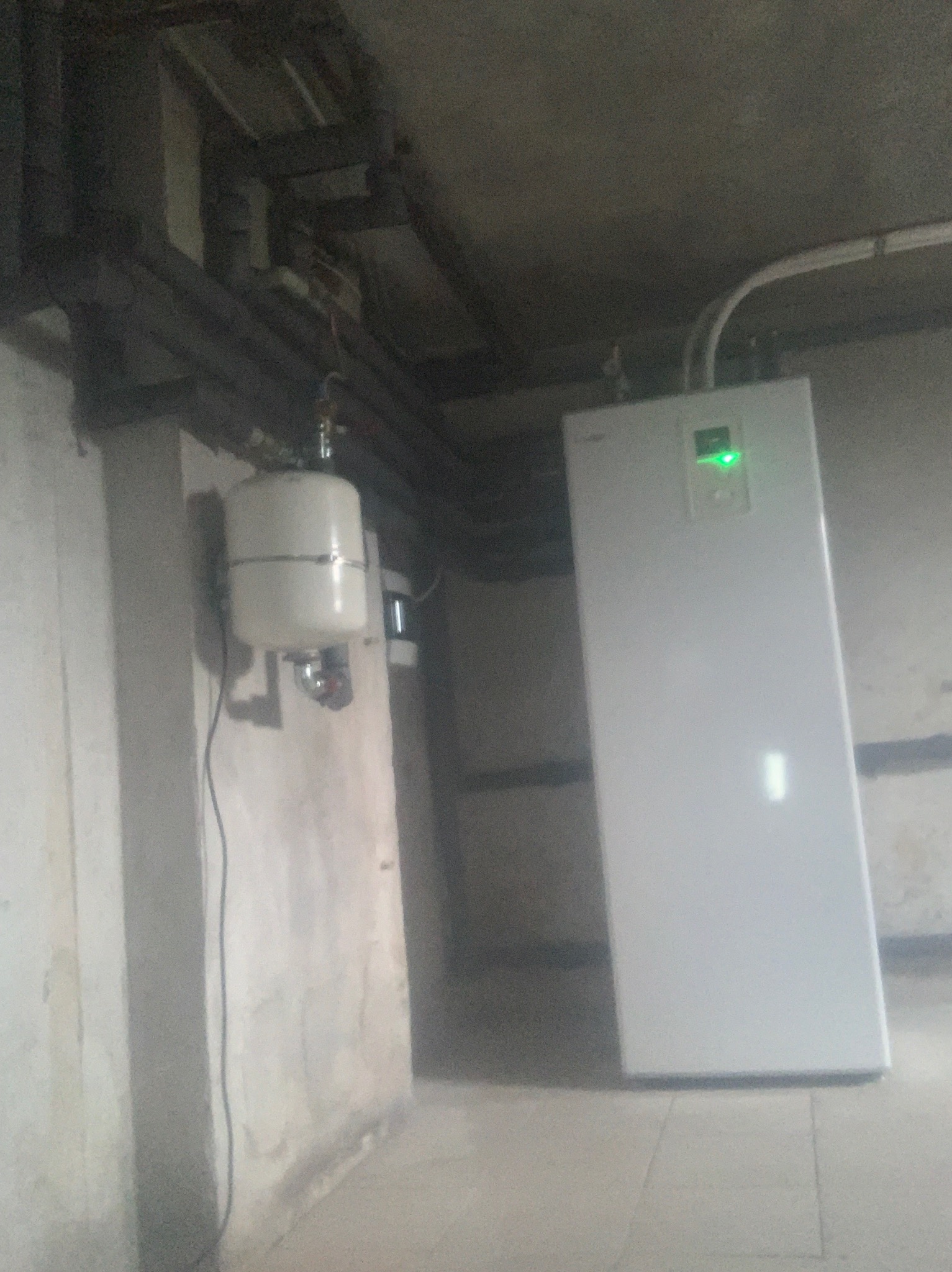 Biała pompa ciepła z zieloną diodą LED na ścianie w piwnicy, widoczne rury instalacyjne i zbiornik wyrównawczy.