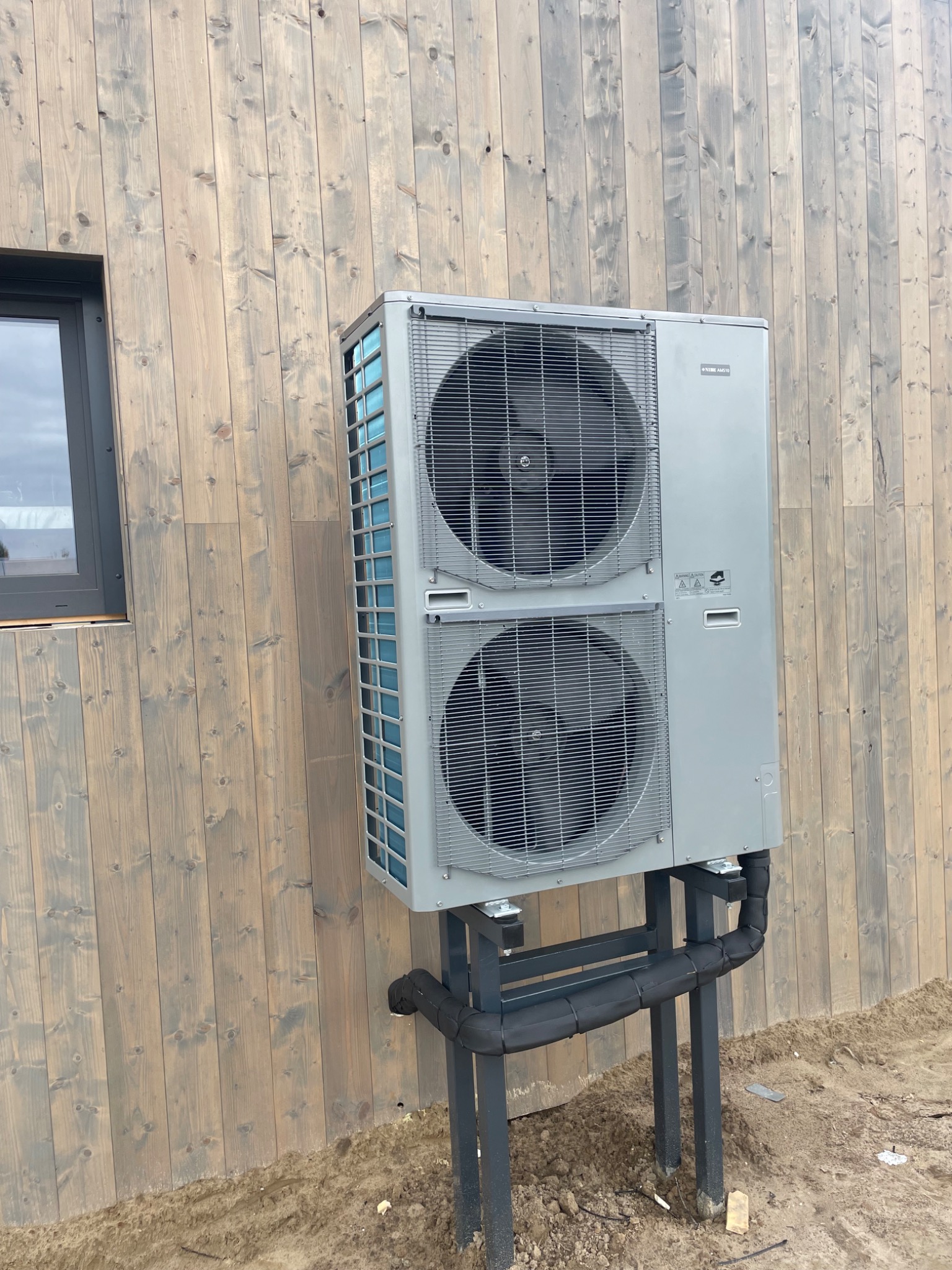 Szara pompa ciepła Nibe AMS10 zamontowana na zewnątrz budynku z drewnianą elewacją. Widoczne rury i elementy instalacji. Ujęcie z boku, blisko okna.