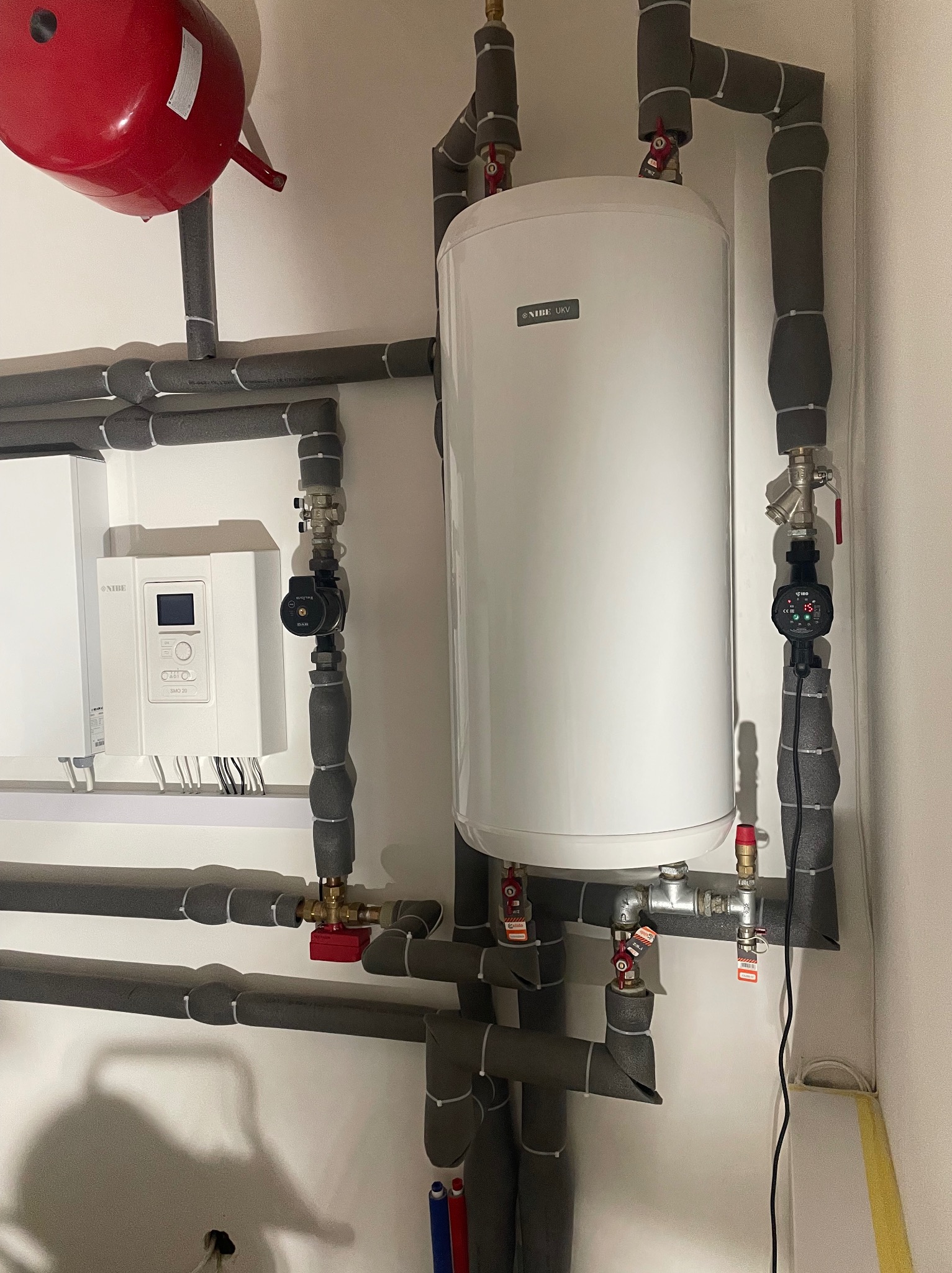 Biały zasobnik ciepłej wody użytkowej Nibe UKV z szarą izolacją rur, zaworami i sterownikiem Nibe SMO 20 na białej ścianie w kotłowni.
