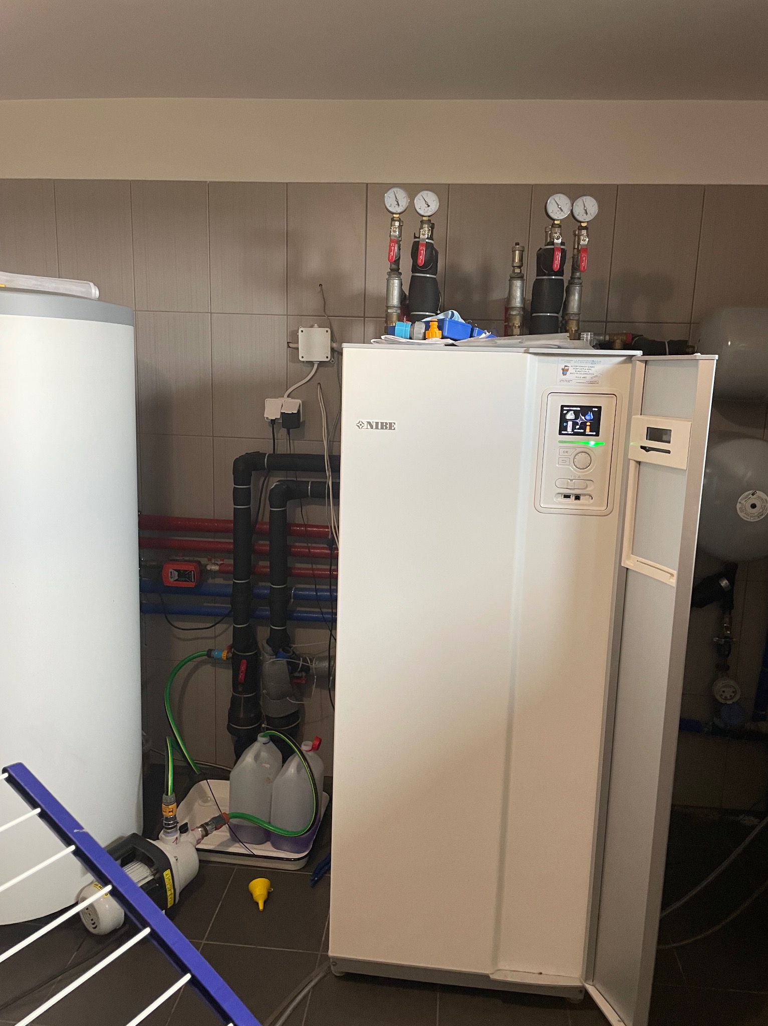 Biała pompa ciepła NIBE z podłączonymi rurami i manometrami, widoczna w kotłowni na tle płytek. Obok zbiornik wyrównawczy i akcesoria instalacyjne.