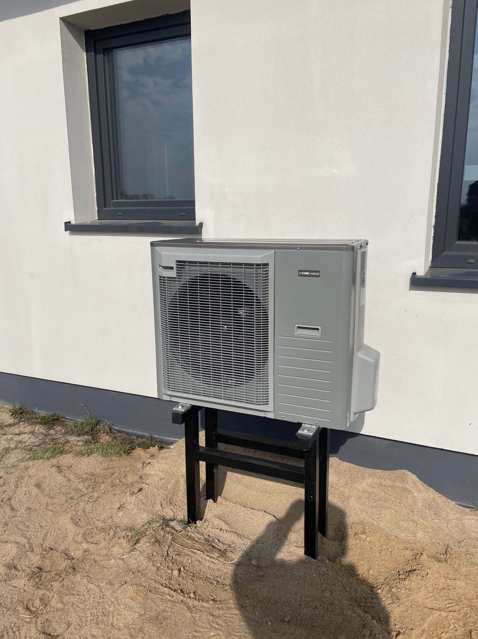 Szara pompa ciepła NIBE AMS20 zamontowana na czarnym stojaku przy białej ścianie budynku z dwoma oknami. Ujęcie z dołu, widoczne podłoże z piasku.