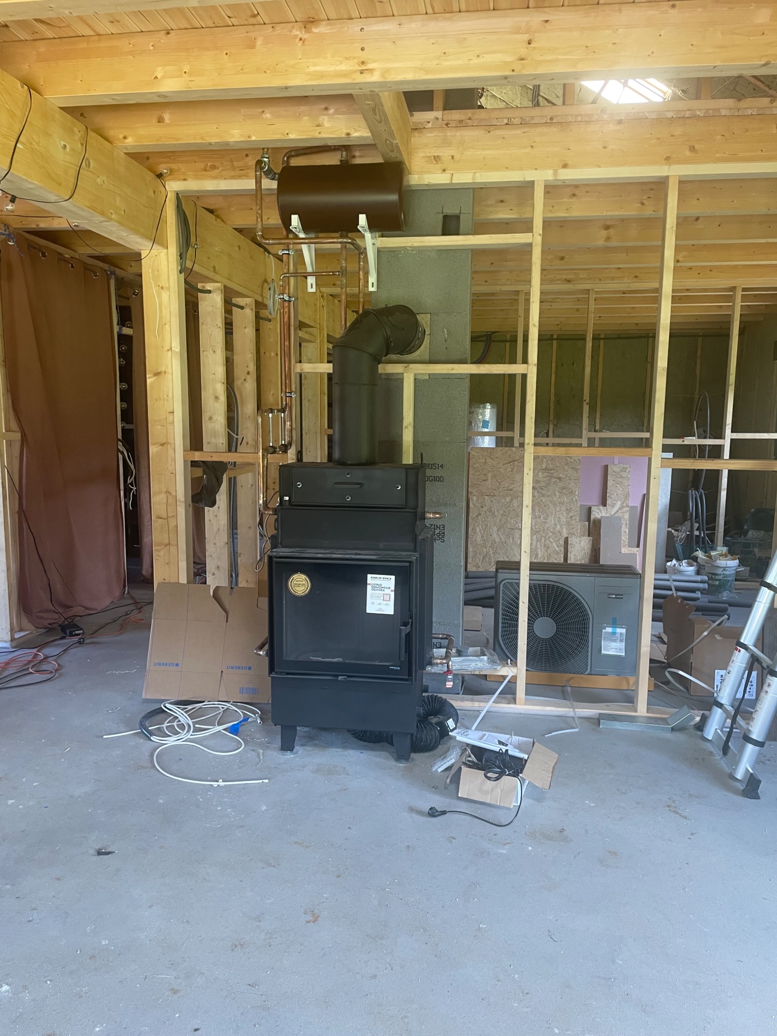 Montaż pompy ciepła i pieca w surowym wnętrzu. Widoczne rury, elementy instalacji grzewczej i klimatyzacji. Częściowo wykończone ściany z drewnianą konstrukcją.