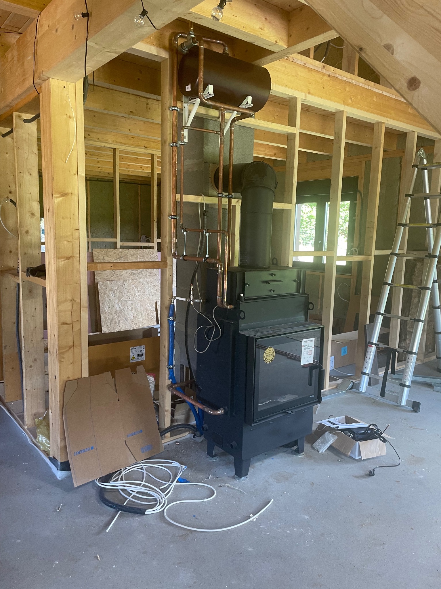 Czarny piec z miedzianą instalacją grzewczą w surowym wnętrzu z drewnianą konstrukcją szkieletową i drabiną. Widoczne elementy instalacyjne na ścianie.