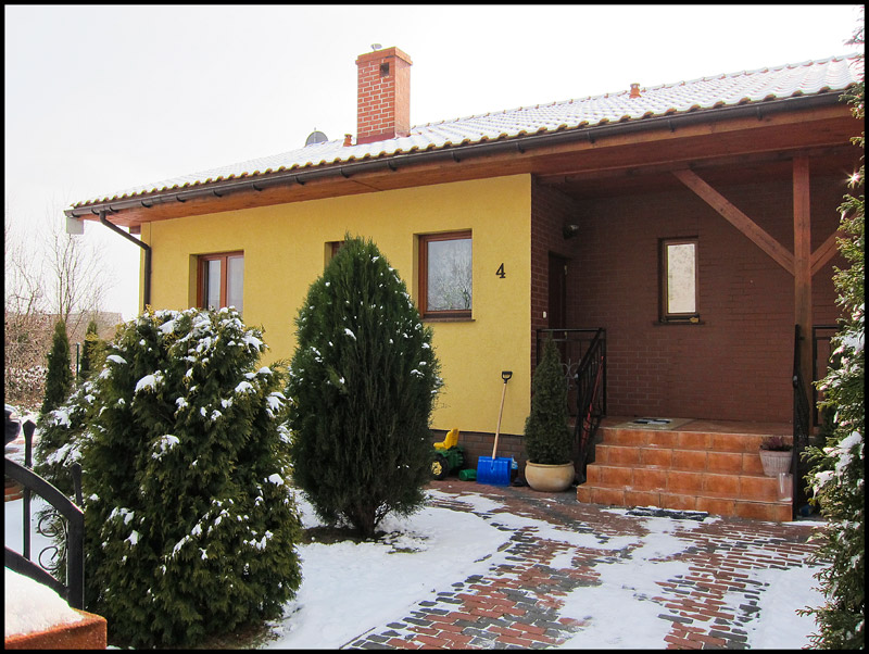 Żółty dom jednorodzinny z ceglanym gankiem i numerem 4, częściowo pokryty śniegiem, widok z zewnątrz w pochmurny dzień.