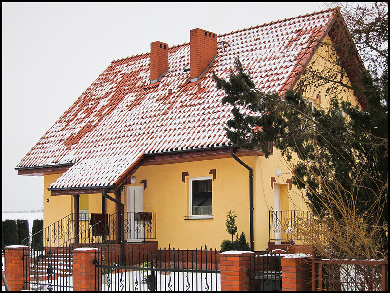 Żółty dom jednorodzinny z czerwonym dachem pokrytym częściowo śniegiem, widoczny zza metalowego ogrodzenia z ceglanymi słupkami.