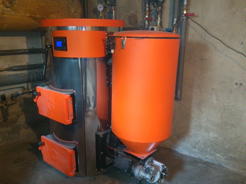 Nowoczesny piec na pellet z pomarańczowym podajnikiem i srebrną obudową, widoczny w kotłowni z betonową ścianą i instalacją rurową.