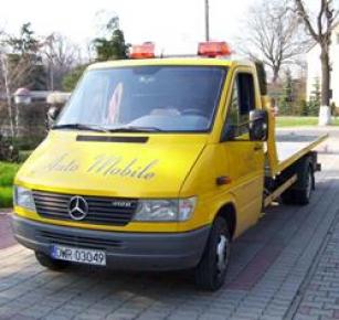 Żółty samochód pomocy drogowej Mercedes Sprinter z napisem 'Auto Mobile' na masce, zaparkowany na chodniku z kostki, z włączonymi pomarańczowymi światłami ostrzegawczymi na dachu.