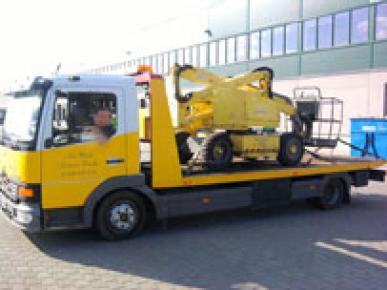 Żółto-biała laweta z żółtym podnośnikiem koszowym na platformie, zaparkowana na tle budynku z licznymi oknami.