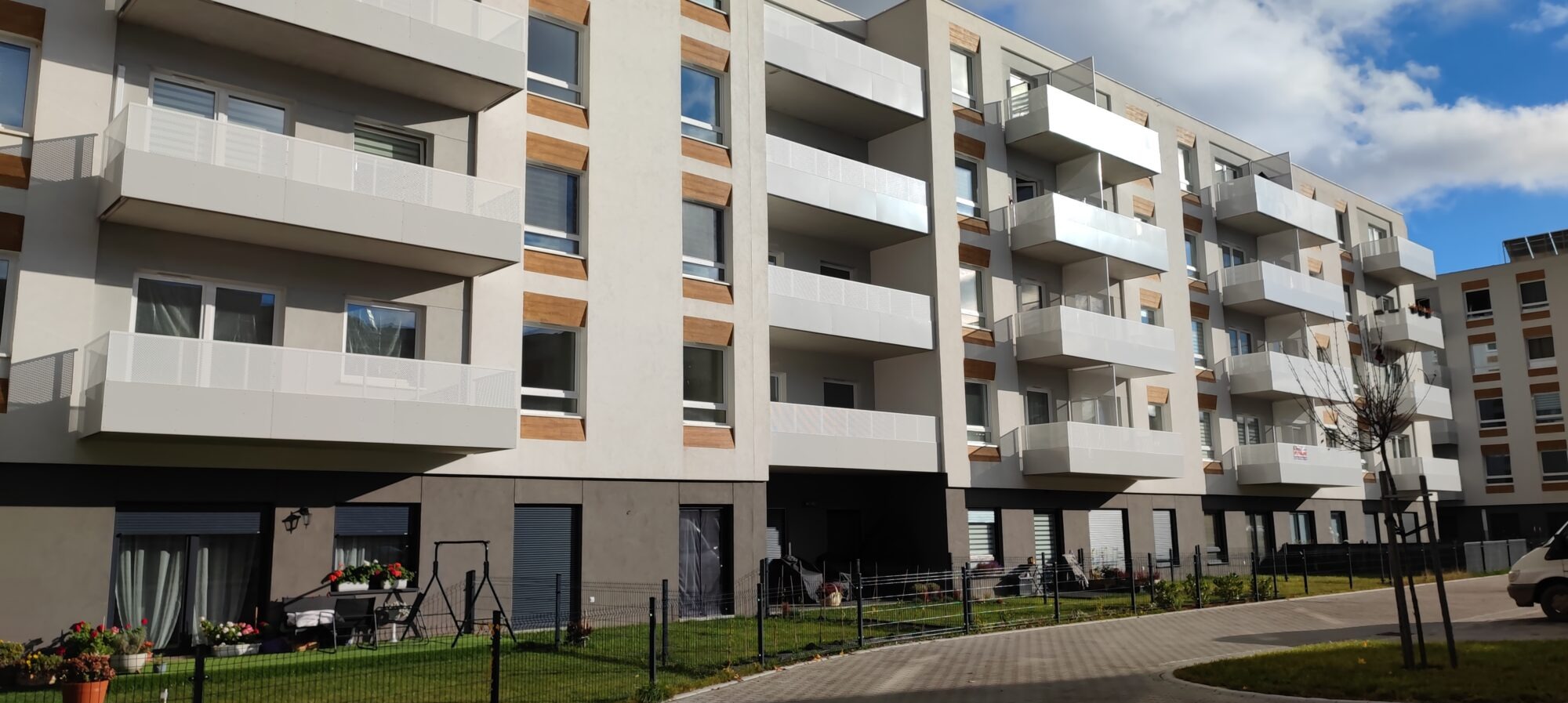 Nowoczesny budynek wielorodzinny z balkonami zabezpieczonymi białymi, ażurowymi balustradami. Widok z poziomu chodnika, słoneczny dzień.