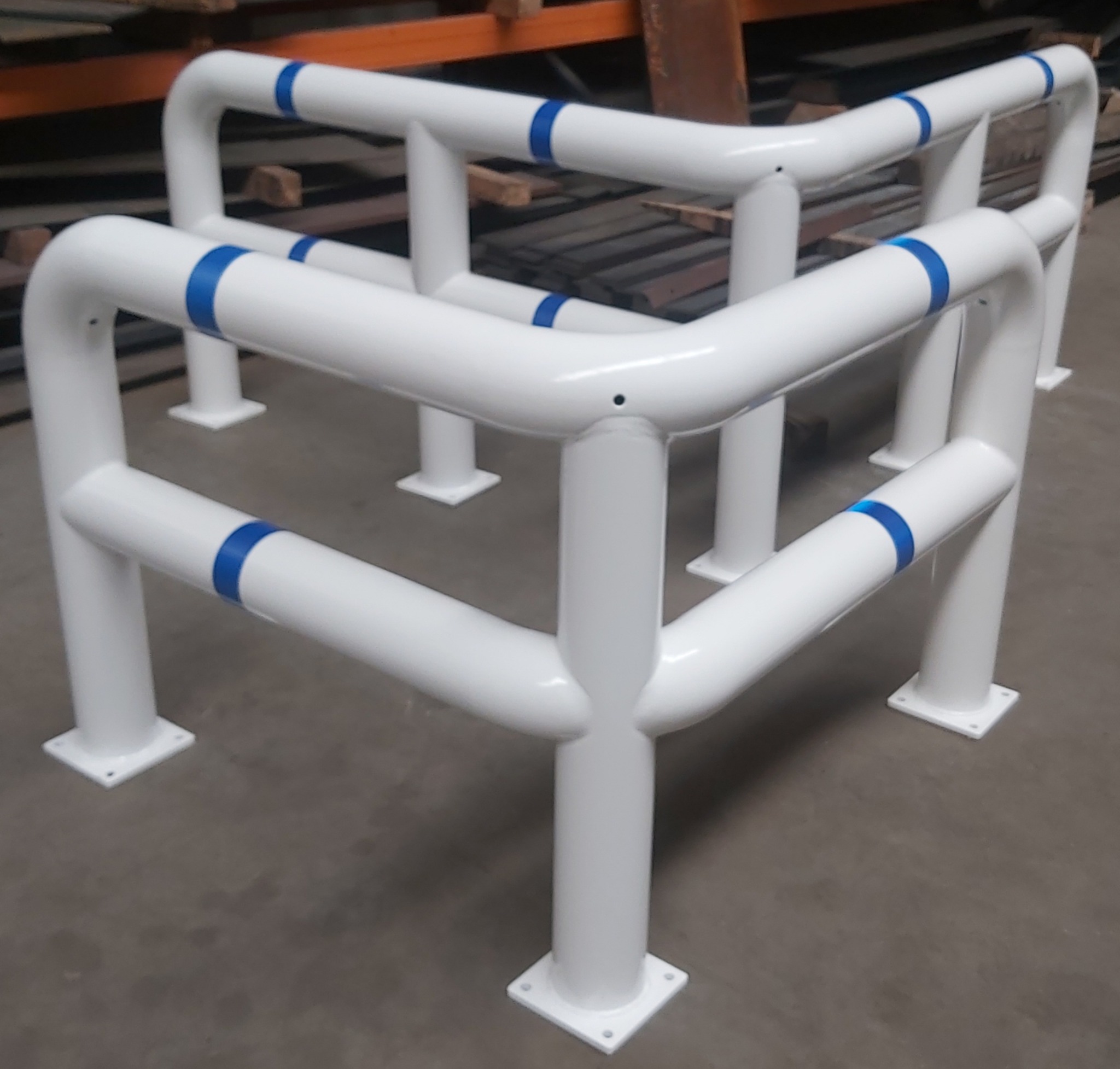 Biała, metalowa odbojnica z niebieskimi akcentami, spawana z rur, ustawiona na szarym podłożu. Widoczne elementy mocujące do podłoża.