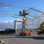 Montaż konstrukcji stalowej hali z użyciem podnośnika nożycowego, widoczne elementy szkieletu dachu oraz pracownicy na platformie, w tle rurociąg i błękitne niebo.