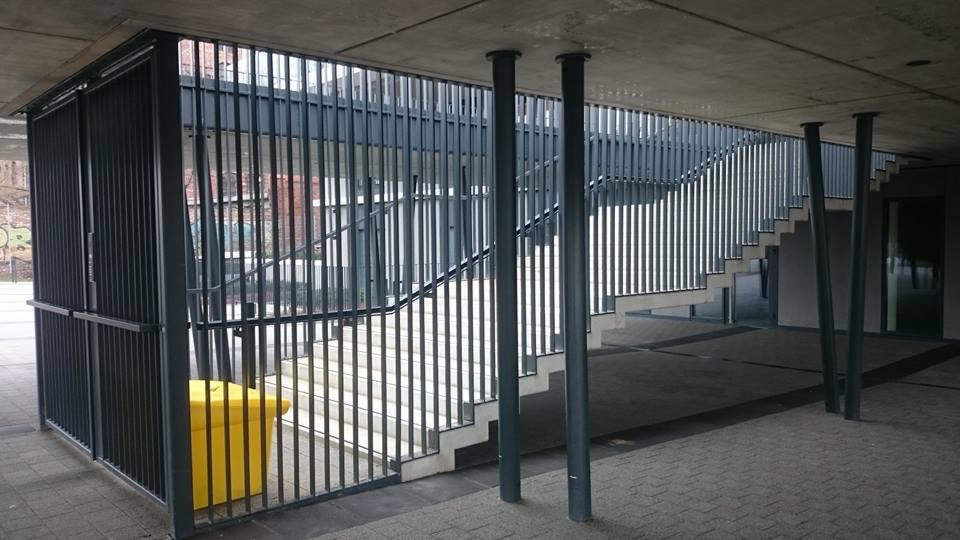 Metalowa barierka z pionowych prętów zabezpieczająca schody zewnętrzne, z żółtym pojemnikiem na śmieci po lewej stronie, widok z dołu z betonowym stropem i filarami.