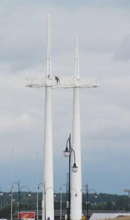 Biała konstrukcja stalowa z platformą widokową, na której znajduje się osoba, widoczna z oddali, na tle pochmurnego nieba i miejskiego krajobrazu z latarniami.