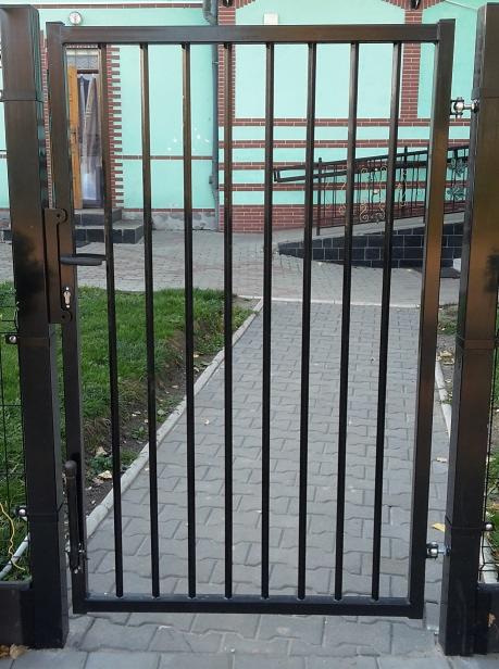 Ciemna, metalowa furtka panelowa z pionowymi szczeblami, widoczna z bliska, z budynkiem w tle i kostką brukową pod spodem.