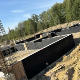 Wykopane i przygotowane fundamenty pod budynek z widocznym zbrojeniem, izolacją pionową i warstwami bloczków betonowych, otoczone ziemią i drzewami w tle.