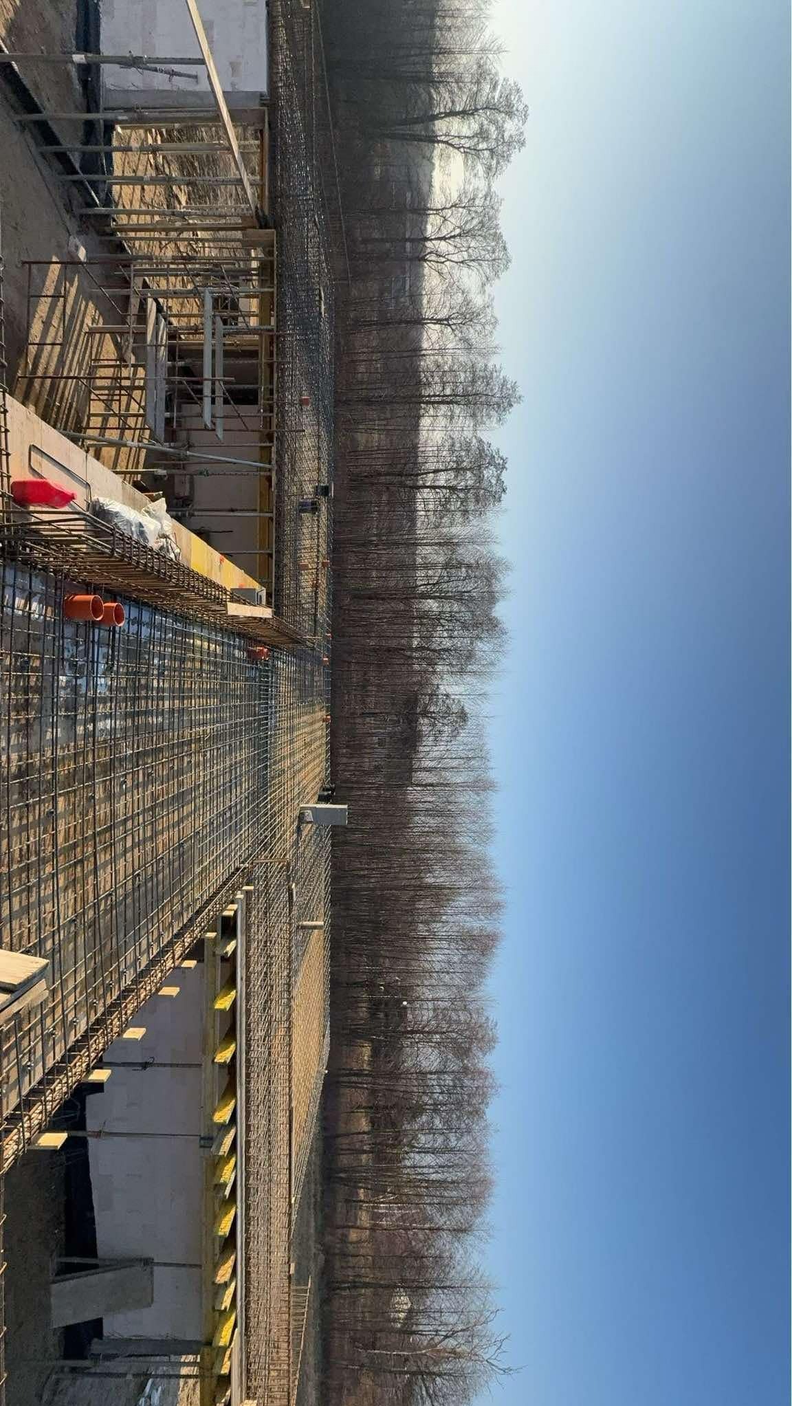 Zbrojenie fundamentów pod budowę w Warszawie, widoczne szalunki i siatka zbrojeniowa na tle drzew.