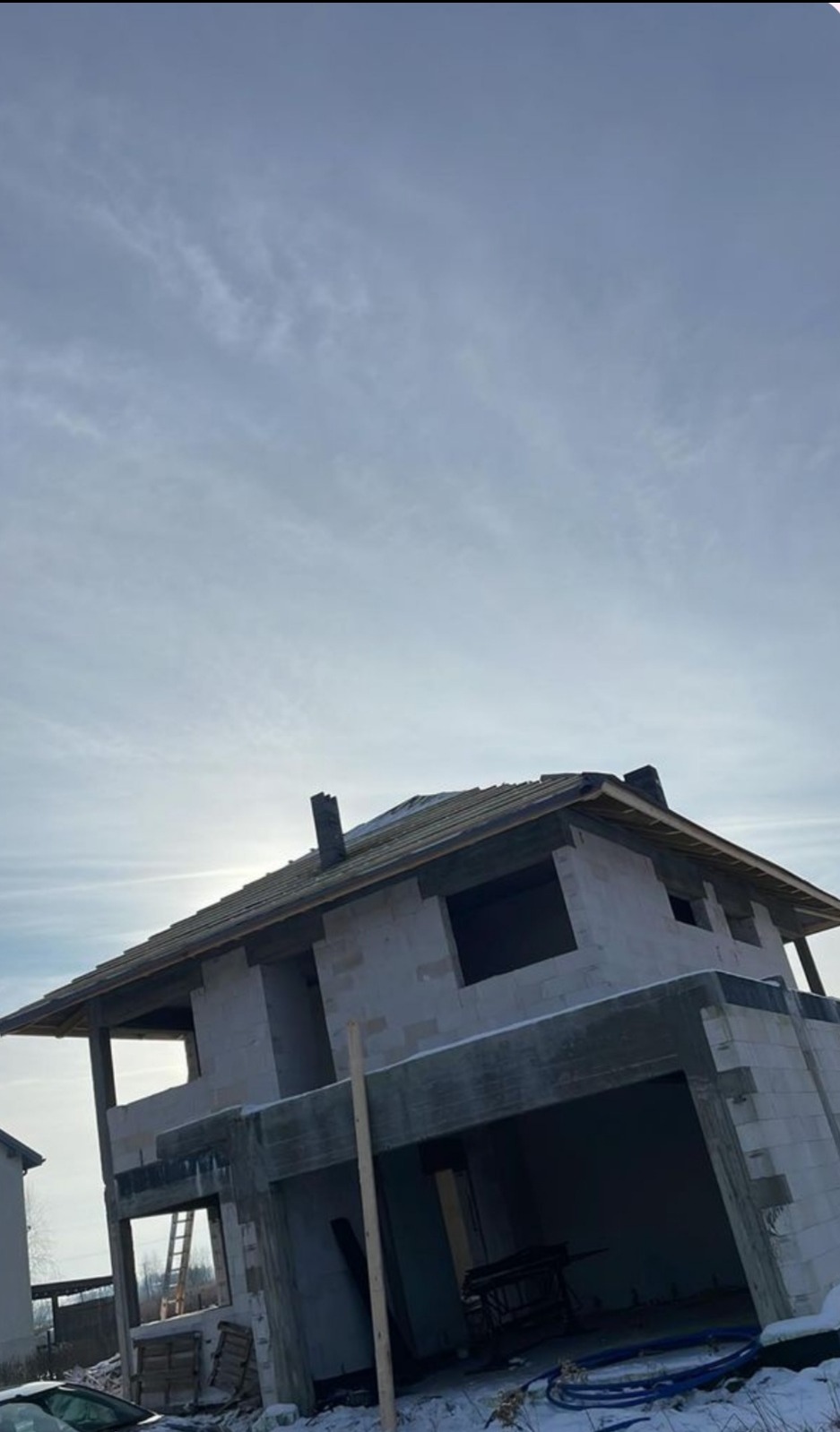 Niedokończony dom z betonu komórkowego w stanie surowym otwartym, widoczne zarysowane okna, dach i komin, w tle pochmurne niebo.