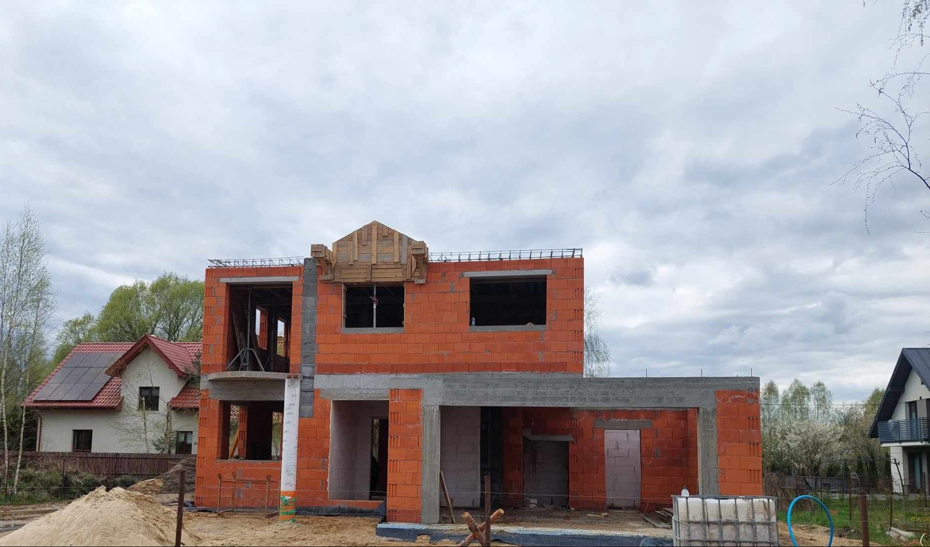 Surowy stan dwukondygnacyjnego domu z czerwonej cegły z widocznym zbrojeniem na dachu i drewnianą konstrukcją nad oknem, na tle pochmurnego nieba i sąsiednich zabudowań.