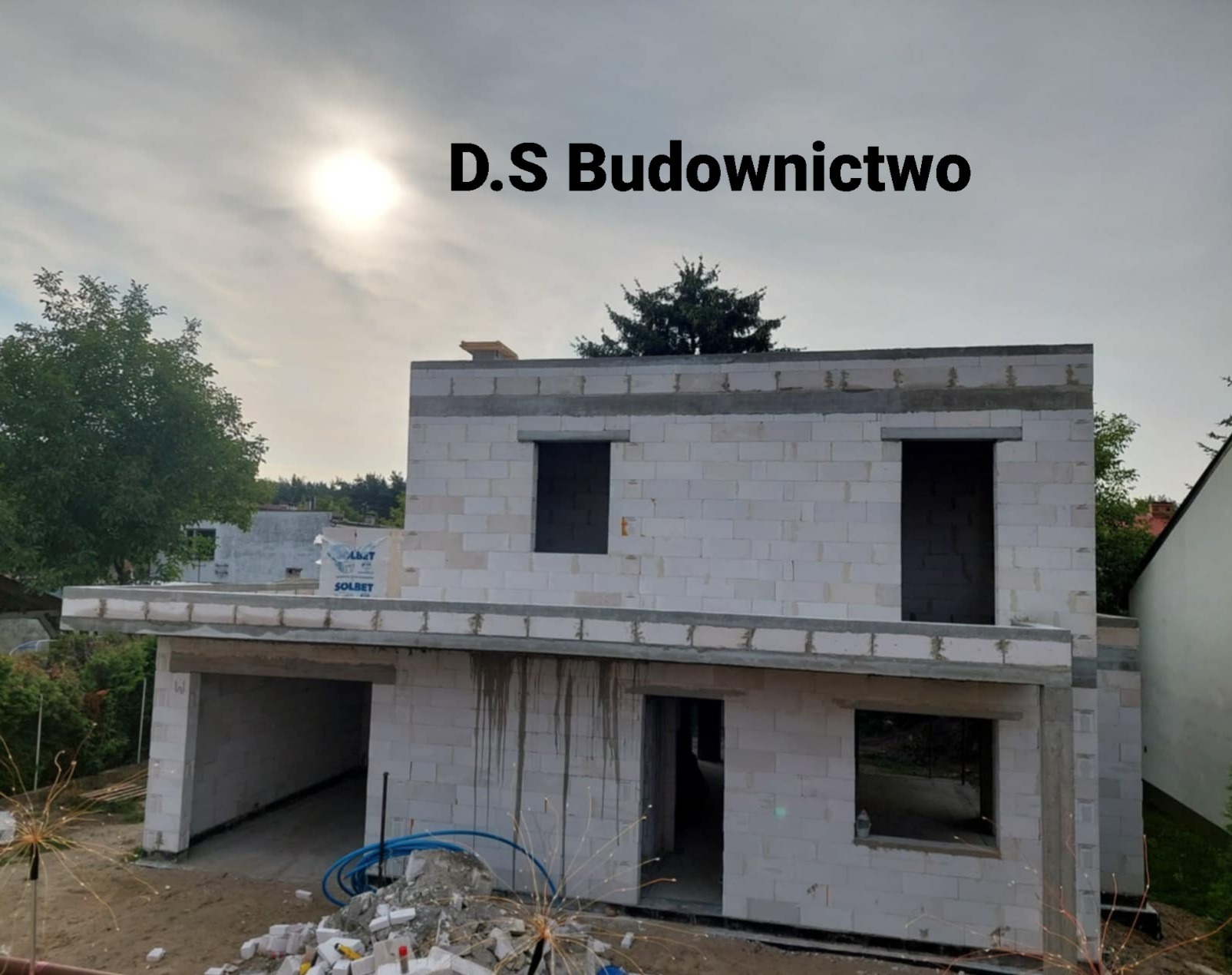 Wznoszony dwukondygnacyjny budynek z betonu komórkowego, widoczne niezabezpieczone otwory okienne, niebieskie rury na ziemi i logo firmy budowlanej w górnej części kadru.